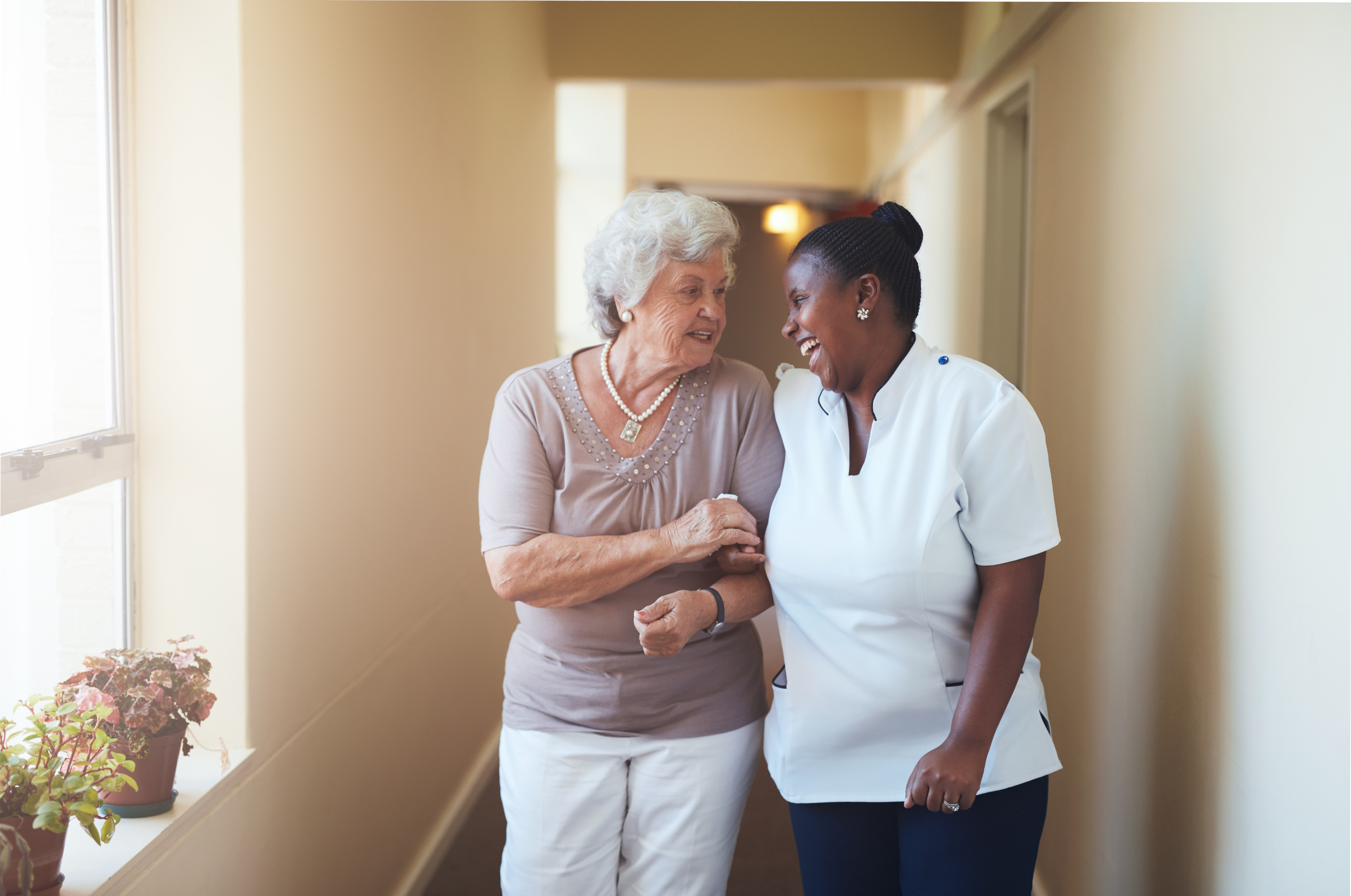 A black woman caregiver assisting and happily conversing with a white elderly woman while walking.